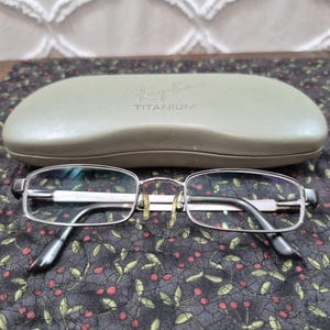 K&ouml;nnte beinhalten: Eine rechteckige Brille aus silberfarbenem Metall mit klaren Gl&auml;sern und goldfarbenen Nasenpads, platziert vor einem hellen olivgr&uuml;nen Hartschalenetui mit dem eingepr&auml;gten Text Ray-Ban und TITANIUM auf der Oberseite.