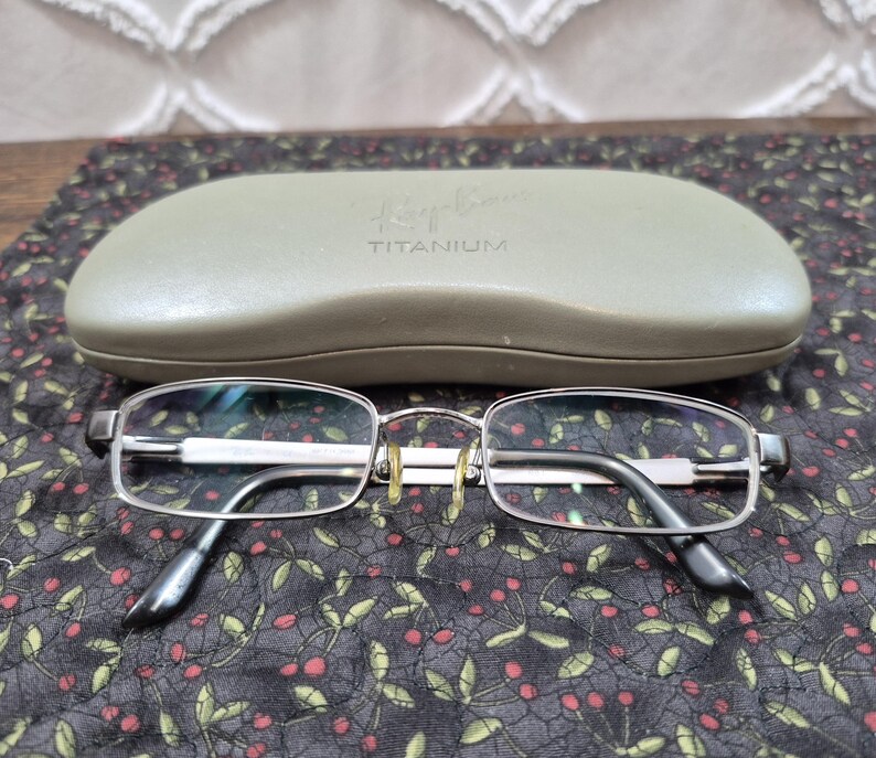 K&ouml;nnte beinhalten: Eine rechteckige Brille aus silberfarbenem Metall mit klaren Gl&auml;sern und goldfarbenen Nasenpads, platziert vor einem hellen olivgr&uuml;nen Hartschalenetui mit dem eingepr&auml;gten Text Ray-Ban und TITANIUM auf der Oberseite.
