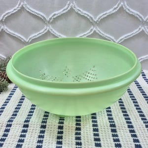 Puede incluir: Un colador de color verde pastel claro con un borde ancho y redondeado y una serie de pequeños orificios de drenaje circulares dispuestos en un patrón decorativo en la base del cuenco. El utensilio de cocina tiene un acabado mate liso.