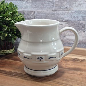 Może przedstawiać: Biały ceramiczny dzbanek z zakrzywionym uchwytem i wylewką. Dzbanek ma wzór plecionki z niebieskimi kwiatowymi akcentami i niebieskim paskiem wokół podstawy. Dzbanek stoi na drewnianej tacy.