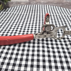 May include: A manual can opener featuring a bright red plastic handle and a metallic silver steel cutting mechanism. The tool includes a rotating gear, a sharp cutting blade, and a bottle opener hook integrated into the metal frame construction.