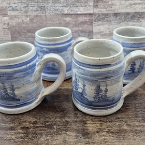 May include: A set of four ceramic mugs with a speckled off-white finish and blue hand-painted horizontal bands featuring pine tree silhouettes. Each mug has a sturdy handle and a cylindrical body, showcasing a consistent rustic design across the collection.