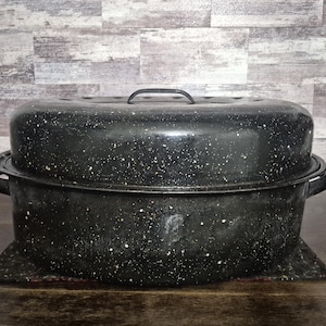May include: A black speckled enamel roasting pan with a lid and handles. The pan sits on a dark metal trivet. The background is a light gray and brown wood-look wall.
