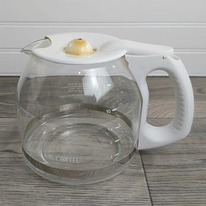 以下が含まれることがあります： 白いハンドルと蓋が付いた、透明なガラス製のMr. Coffeeコーヒーカラフ。カラフには12カップまでの目盛りが付いており、中央にはシルバーのバンドが付いています。ハンドルは湾曲しており、蓋には小さなタン色のノブが付いています。