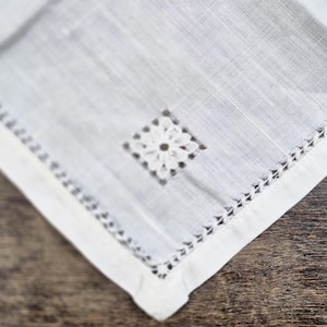 May include: Close-up of a white linen handkerchief with a delicate floral embroidered square in the center. The edges are finished with a decorative stitched border. The fabric has a subtle texture.