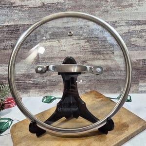 Op de afbeelding: Een heldere glazen Masterclass deksel met een roestvrijstalen rand en handvat. Het deksel wordt ondersteund door een zwarte standaard, rustend op een houten snijplank. De deksel is ontworpen voor kookgerei.