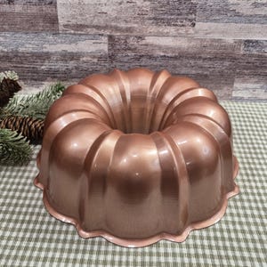 May include: A copper-colored bundt cake pan with a fluted design. The pan has a rounded shape with a central hole and a scalloped edge. The pan is made of metal and has a shiny finish. The pan is sitting on a green and white checkered surface.