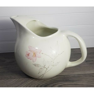 May include: Cream ceramic pitcher with a round body and a curved handle. A pink floral design with green stems and leaves is painted on the front. The pitcher has a spout for pouring.