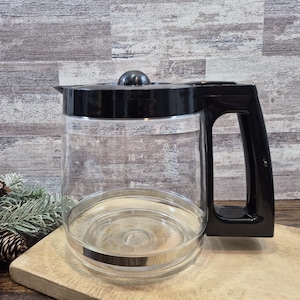 May include: A clear glass coffee carafe featuring a black plastic lid and a matching black handle. The glass body displays printed measurement markings for 8 and 10 cups, along with a warning symbol and text indicating caution for hot surfaces.