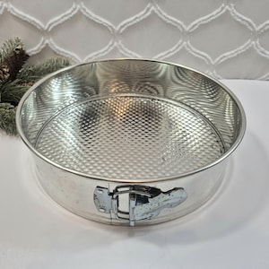 May include: A round silver-colored metal springform cake pan with a textured diamond pattern on the base. The pan features a side latch mechanism for releasing the outer ring, constructed from polished steel with a reflective surface finish.
