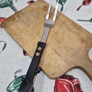 May include: A vintage carving fork with a black handle and brass rivets, resting on a wooden cutting board. The fork has two tines and a stainless steel blade. The cutting board is rectangular with a handle.
