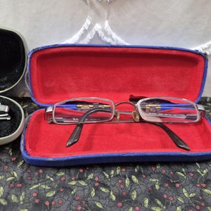 K&ouml;nnte beinhalten: Silberfarbene Metallbrille mit klaren Gl&auml;sern in einem rechteckigen, blauen Hartschalenetui mit leuchtend rotem Innenfutter. Die Brille hat einen d&uuml;nnen Rahmen und dunkle B&uuml;gelenden, wobei das Ray-Ban-Logo auf dem Glas sichtbar ist.