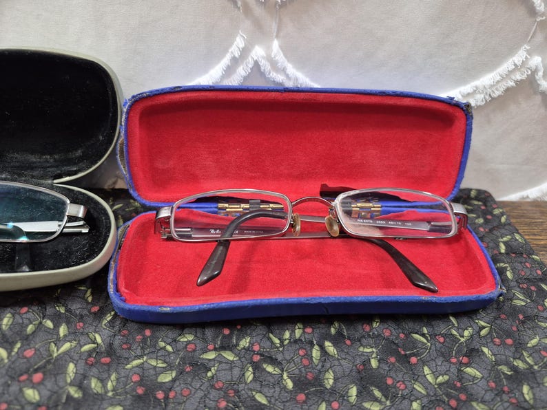 K&ouml;nnte beinhalten: Silberfarbene Metallbrille mit klaren Gl&auml;sern in einem rechteckigen, blauen Hartschalenetui mit leuchtend rotem Innenfutter. Die Brille hat einen d&uuml;nnen Rahmen und dunkle B&uuml;gelenden, wobei das Ray-Ban-Logo auf dem Glas sichtbar ist.