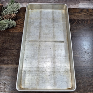 May include: A rectangular, well-worn, aluminum baking sheet with a single horizontal groove. The sheet has a slightly raised edge and shows signs of use. The background features a wooden surface and decorative pinecones.