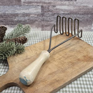 May include: A vintage potato masher with a light wood handle and a metal mashing head. The masher is resting on a wooden cutting board. The handle has a light blue paint on the bottom.