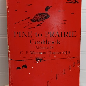 May include: A vintage cookbook titled "Pine to Prairie Cookbook" with a red cover. The cover features a black illustration of a loon, trees, and text. The book is Volume IV, Chapter #18, by C. P. Wainman.