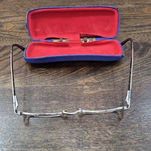 K&ouml;nnte beinhalten: Eine Brille mit d&uuml;nnem Metallrahmen und ein blaues Hartschalenetui mit leuchtend rotem Innenfutter. Die Brille ist vor dem ge&ouml;ffneten Etui positioniert und zeigt deutlich den Steg, die Nasenpads und die d&uuml;nnen B&uuml;gel.