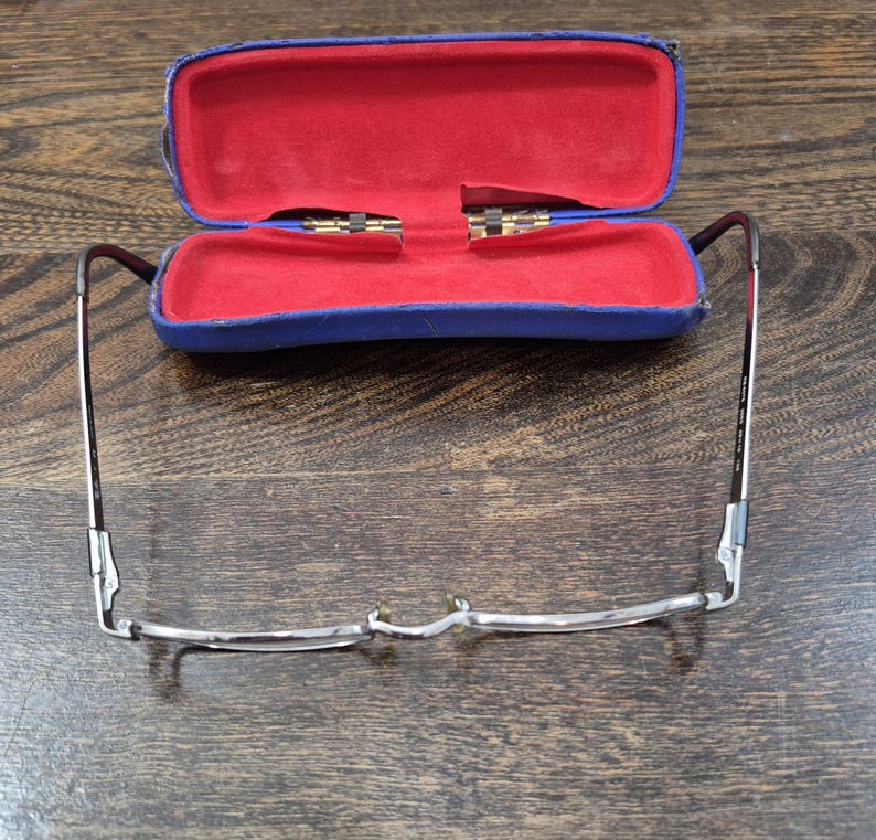 K&ouml;nnte beinhalten: Eine Brille mit d&uuml;nnem Metallrahmen und ein blaues Hartschalenetui mit leuchtend rotem Innenfutter. Die Brille ist vor dem ge&ouml;ffneten Etui positioniert und zeigt deutlich den Steg, die Nasenpads und die d&uuml;nnen B&uuml;gel.
