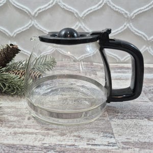 Può includere: Una caraffa per caffè in vetro trasparente con coperchio in plastica nera e una robusta impugnatura nera. La caraffa presenta una base ampia e arrotondata e una parte superiore affusolata. La parola MAX è stampata in piccole lettere bianche.