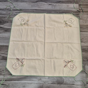 Puede incluir: Delantal color crema con ribete y lazos verde claro. El delantal presenta bordados de palos de cartas y pájaros. Los palos son corazones, picas, diamantes y tréboles. El delantal está hecho de una tela suave.