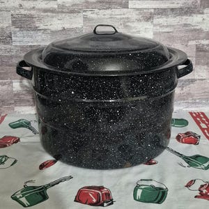 May include: A large, black speckled enamelware pot with a lid and two side handles. The pot is round and has a wide rim. The lid has a small handle. The pot is sitting on a patterned tablecloth.