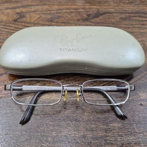 K&ouml;nnte beinhalten: Ray-Ban Titanbrille mit einem d&uuml;nnen, rechteckigen, schie&szlig;grauen Rahmen und klaren Gl&auml;sern. Die Brille verf&uuml;gt &uuml;ber gelbe Nasenpads und schwarze B&uuml;gelenden. Ein hellgraues, hartes Schutzetui mit dem Ray-Ban-Logo und dem eingepr&auml;gten Wort TITANIUM auf der Oberseite ist enthalten.