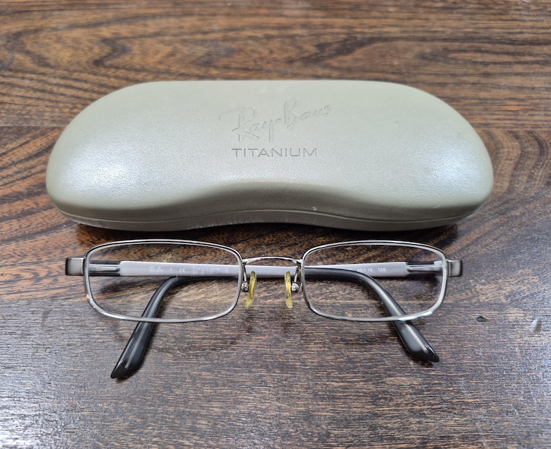 K&ouml;nnte beinhalten: Ray-Ban Titanbrille mit einem d&uuml;nnen, rechteckigen, schie&szlig;grauen Rahmen und klaren Gl&auml;sern. Die Brille verf&uuml;gt &uuml;ber gelbe Nasenpads und schwarze B&uuml;gelenden. Ein hellgraues, hartes Schutzetui mit dem Ray-Ban-Logo und dem eingepr&auml;gten Wort TITANIUM auf der Oberseite ist enthalten.