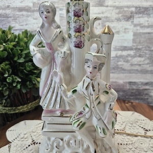 Może przedstawiać: Zabytkowa lampa porcelanowa z dwiema postaciami w strojach z epoki. Podstawa lampy jest biała ze złotymi akcentami i kwiatowym wzorem. Postacie są pomalowane z delikatnymi detalami, a lampa ma mosiężny wierzch.