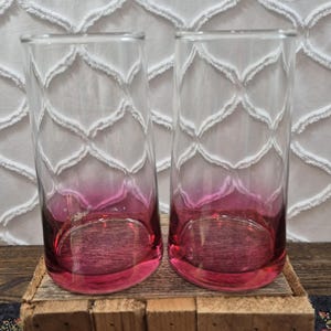 Könnte beinhalten: Zwei hohe, zylindrische Trinkgläser aus klarem Glas. Jedes Glas weist einen ausgeprägten Ombré-Effekt auf, der von einem tiefen, gesättigten Magentarot am schweren Boden zu einem völlig transparenten, farblosen oberen Rand übergeht.