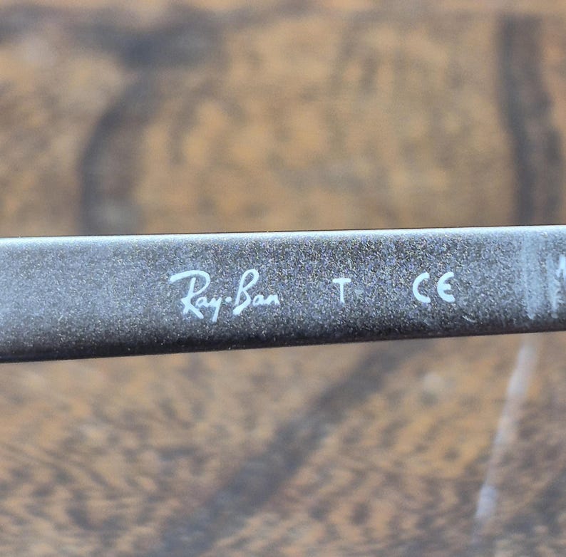 K&ouml;nnte beinhalten: Ein dunkelgrauer oder schwarzer Brillenb&uuml;gel mit dem wei&szlig;en Aufdruck Ray-Ban, gefolgt von dem Buchstaben T und dem CE-Zeichen. Die Oberfl&auml;che des B&uuml;gels hat ein leicht texturiertes, mattes Finish mit sichtbaren hellen Staubpartikeln.