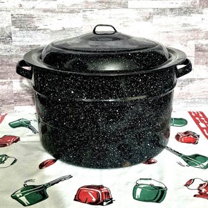 May include: A large black stockpot with a speckled white finish, featuring two side handles and a matching lid with a central handle. The pot has a cylindrical shape with a stepped design along the sides, constructed from enameled metal.