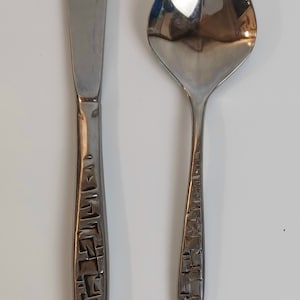May include: A stainless steel butter knife and spoon set. The knife has a flat, rounded blade. Both utensils have textured handles with a geometric pattern. The spoon has a rounded bowl and a long, slender handle.