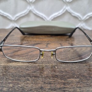 K&ouml;nnte beinhalten: Rechteckige Brille mit einem d&uuml;nnen, dunkelmetallischen Rahmen und klaren Gl&auml;sern. Die Brille verf&uuml;gt &uuml;ber verstellbare Nasenpads mit einem gelblichen Schimmer und schmale B&uuml;gel. Ein passendes, helles Hartschalenetui befindet sich hinter dem Rahmen.