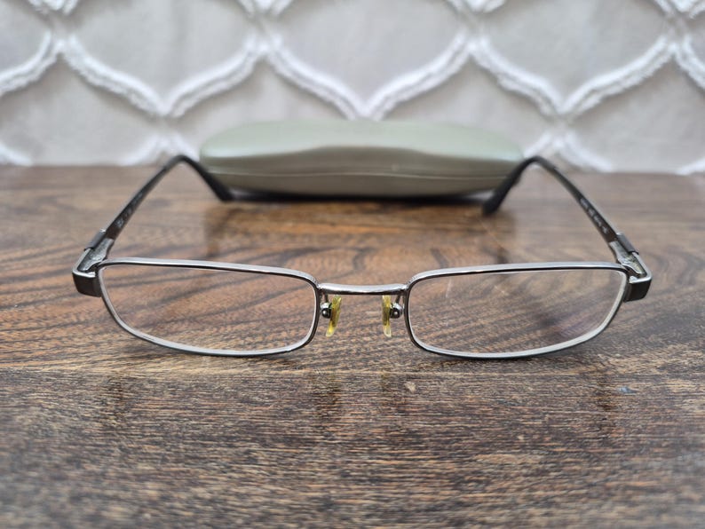 K&ouml;nnte beinhalten: Rechteckige Brille mit einem d&uuml;nnen, dunkelmetallischen Rahmen und klaren Gl&auml;sern. Die Brille verf&uuml;gt &uuml;ber verstellbare Nasenpads mit einem gelblichen Schimmer und schmale B&uuml;gel. Ein passendes, helles Hartschalenetui befindet sich hinter dem Rahmen.