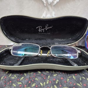 K&ouml;nnte beinhalten: Silberfarbene rechteckige Brille mit klaren Gl&auml;sern und gelben Nasenpads, die in einem harten, olivgr&uuml;nen Ray-Ban Titanium Etui mit schwarzem Innenfutter liegt. Der Deckel des Etuis zeigt das wei&szlig;e Ray-Ban Titanium Logo.