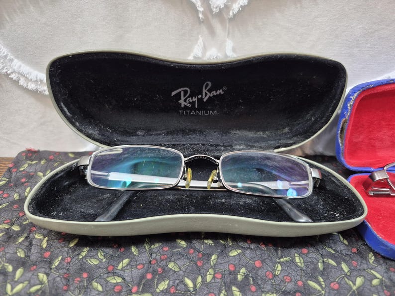 K&ouml;nnte beinhalten: Silberfarbene rechteckige Brille mit klaren Gl&auml;sern und gelben Nasenpads, die in einem harten, olivgr&uuml;nen Ray-Ban Titanium Etui mit schwarzem Innenfutter liegt. Der Deckel des Etuis zeigt das wei&szlig;e Ray-Ban Titanium Logo.