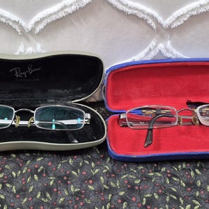 K&ouml;nnte beinhalten: Zwei Brillen mit rechteckigen Gl&auml;sern und Metallrahmen. Ein Paar liegt in einem grauen Etui mit dem aufgedruckten Ray-Ban Titanium Logo. Das zweite Paar befindet sich in einem blauen Etui mit leuchtend rotem Innenfutter. Beide Etuis sind offen.