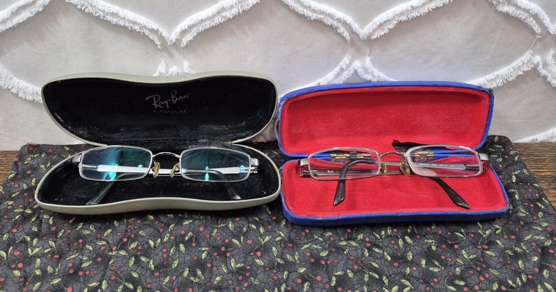 K&ouml;nnte beinhalten: Zwei Brillen mit rechteckigen Gl&auml;sern und Metallrahmen. Ein Paar liegt in einem grauen Etui mit dem aufgedruckten Ray-Ban Titanium Logo. Das zweite Paar befindet sich in einem blauen Etui mit leuchtend rotem Innenfutter. Beide Etuis sind offen.