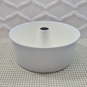 May include: A solid white round baking pan with a smooth finish and a central tube, designed for ring-shaped cakes. The pan features a wide, open top and a flat base, with a dark grey interior rim visible at the very top of the central chimney.