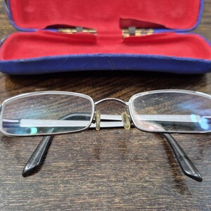 K&ouml;nnte beinhalten: Eine rechteckige Brille mit silberfarbenem Metallrahmen und schwarzen B&uuml;geln, ausgestattet mit klaren Gl&auml;sern und vergilbten Nasenpads, platziert vor einem ge&ouml;ffneten blauen Schutzetui mit leuchtend rotem Innenfutter.