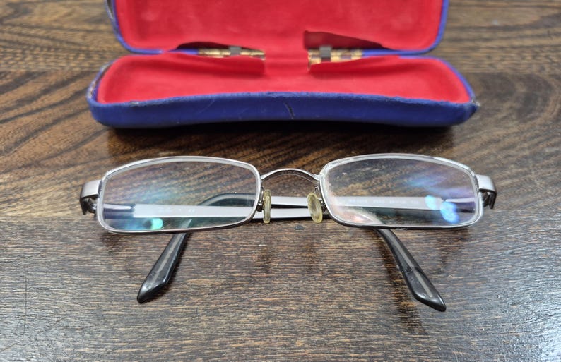 K&ouml;nnte beinhalten: Eine rechteckige Brille mit silberfarbenem Metallrahmen und schwarzen B&uuml;geln, ausgestattet mit klaren Gl&auml;sern und vergilbten Nasenpads, platziert vor einem ge&ouml;ffneten blauen Schutzetui mit leuchtend rotem Innenfutter.