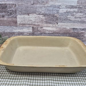 May include: A rectangular, off-white stoneware baking dish with rounded corners and a slightly raised rim. The dish has a vintage appearance, suggesting it is well-used. The background features a rustic wooden wall and a checkered tablecloth.