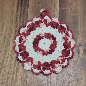 May include: A handmade, round crochet potholder in shades of cream, red, and pink. The potholder features a scalloped edge and a loop for hanging. The design is a circular pattern with concentric rings of color.