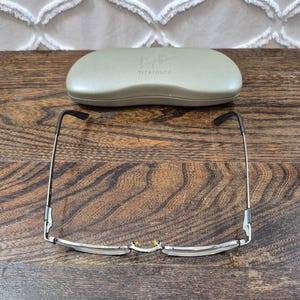 K&ouml;nnte beinhalten: Eine silberfarbene Metallbrille mit d&uuml;nnem Rahmen und schwarzen B&uuml;gelenden, platziert vor einem hellen olivgr&uuml;nen Hartschalenetui. Das Etui ist auf der Oberseite mit dem gepr&auml;gten Schriftzug Ray-Ban und TITANIUM versehen.