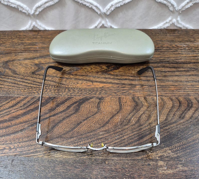 K&ouml;nnte beinhalten: Eine silberfarbene Metallbrille mit d&uuml;nnem Rahmen und schwarzen B&uuml;gelenden, platziert vor einem hellen olivgr&uuml;nen Hartschalenetui. Das Etui ist auf der Oberseite mit dem gepr&auml;gten Schriftzug Ray-Ban und TITANIUM versehen.