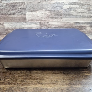 May include: A rectangular food storage container with a navy blue lid and a silver-colored base. The lid features a white apple design. The container is sitting on a wooden surface.