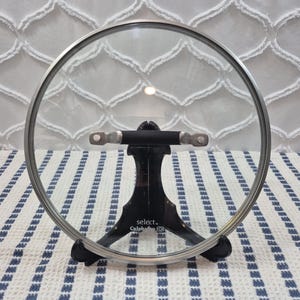 Könnte beinhalten: Ein runder Topfdeckel aus klarem Glas mit einem silberfarbenen Metallrand und einem horizontalen schwarzen Griff. Der Deckel wird von einem schwarzen Kunststoffständer gehalten, auf dessen Vorderseite select by Calphalon in Weiß steht.