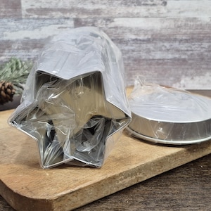 May include: A set of metal cookie cutters, including a star shape and a round shape, packaged in clear plastic. The cutters are displayed on a wooden cutting board, suggesting a baking theme.