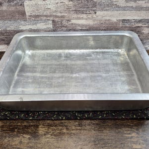 May include: A rectangular, aluminum baking pan with slightly rounded corners. The pan sits on a dark, patterned placemat, against a backdrop of wood paneling. The pan is empty and shows signs of use.