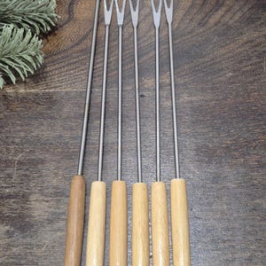 May include: Set of six fondue forks with wooden handles and stainless steel tines. The handles are natural wood with colored tips in blue, yellow, blue, green, red, and gray. The forks are arranged vertically on a dark wood surface.